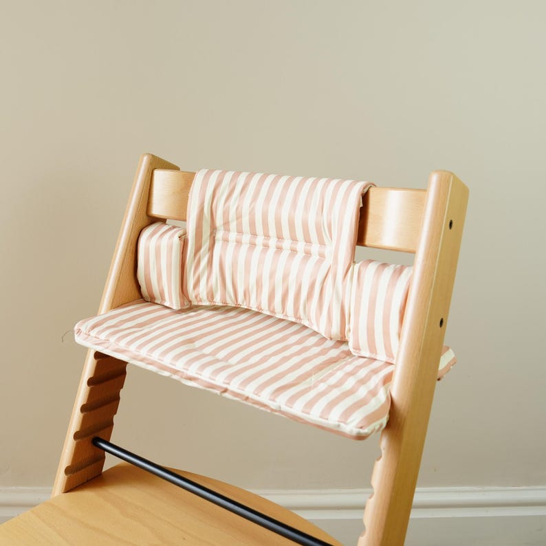 May include: A wooden high chair with a striped cushion set in light pink and white. The chair has a natural wood finish and a black bar for foot support. The cushion set includes a seat cushion, back cushion, and side cushions.