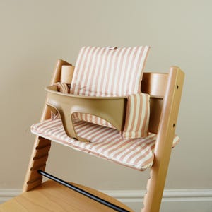 May include: A wooden high chair with a tan plastic tray and striped cushions in shades of pink and white. The chair has a black footrest and is designed for a baby or toddler.