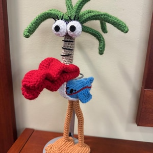 May include: A whimsical, handmade crochet palm tree sculpture. The tree features a green crown, large googly eyes, and a red scarf. The sculpture has long legs and feet, and a blue and white accent. It is mounted on a silver base.