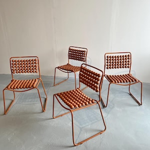 Puede incluir: Cuatro sillas de metal naranja con asientos de cuentas marrones. Las sillas están dispuestas en forma de cuadrado sobre un suelo de hormigón gris.