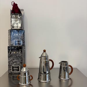 Pode incluir: Uma coleção de máquinas de café expresso em aço inoxidável com alças e botões de madeira. A imagem também mostra várias caixas com o texto "ARMONIE Opera". As máquinas de café expresso estão em uma superfície reflexiva.