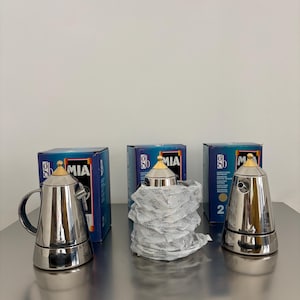 May include: Three silver-coloured metal espresso makers with gold-toned conical knobs on the lids. Two of the espresso makers are fully visible, while one is partially wrapped in white protective paper. Each unit is accompanied by a blue box labelled MIA.