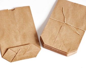 May include: A set of light brown paper bags with a rectangular base and a thumb notch at the top opening. One bag is laid flat showing the folded bottom, while a stack of identical bags is tied together with a thin piece of twine.