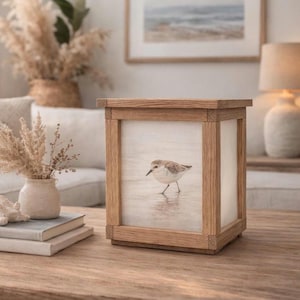 Könnte beinhalten: Eine Holzlaterne mit einem gerahmten Bild eines kleinen Vogels am Strand. Die Laterne hat eine quadratische Form mit einem hellen Holzrahmen. Das Bild ist durch die Vorder- und Seitenwände sichtbar. Die Laterne steht auf einem Holztisch.