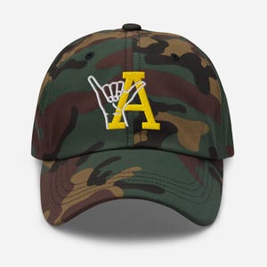May include: A camouflage baseball cap with a yellow embroidered letter 'A' and a white embroidered hand gesture.