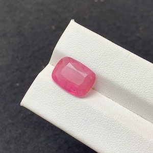 Puede incluir: Una piedra preciosa de corte cojín con un tono rosa vibrante y una superficie facetada. La piedra se muestra sobre un soporte de exhibición blanco, texturizado e inclinado. La piedra tiene un aspecto claro y pulido con reflejos de luz.
