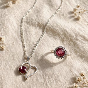 Puede incluir: Un collar de plata con un colgante en forma de corazón que presenta una piedra preciosa redonda de color rosa oscuro y pequeñas piedras claras, junto a un anillo de plata a juego con una piedra rosa oscuro rodeada por un halo de piedras claras.