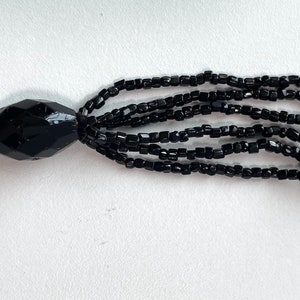 K&ouml;nnte beinhalten: Eine schwarze Perlenkette mit einem Quastenanh&auml;nger. Die Kette besteht aus kleinen, facettierten Perlen und hat eine gro&szlig;e, facettierte Perle am Ende der Quaste.