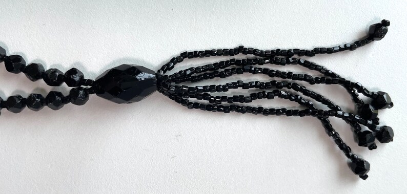 K&ouml;nnte beinhalten: Eine schwarze Perlenkette mit einem Quastenanh&auml;nger. Die Kette besteht aus kleinen, facettierten Perlen und hat eine gro&szlig;e, facettierte Perle am Ende der Quaste.