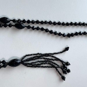 K&ouml;nnte beinhalten: Eine schwarze Perlenkette mit einem Quastenanh&auml;nger. Die Kette besteht aus kleinen, runden Perlen und hat einen langen, herabh&auml;ngenden Quastenanh&auml;nger am Ende.