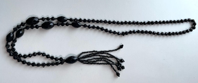 K&ouml;nnte beinhalten: Eine schwarze Perlenkette mit einem Quastenanh&auml;nger. Die Kette besteht aus kleinen, runden Perlen und hat einen langen, herabh&auml;ngenden Quastenanh&auml;nger am Ende.