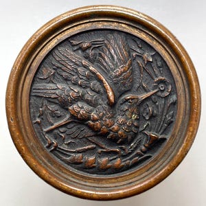 May include: A round, bronze-colored object with a detailed relief carving of a hummingbird in flight, surrounded by foliage. The bird is depicted with outstretched wings and a long beak, with a detailed border.