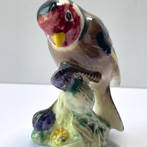 May include: A ceramic figurine of a bird perched on a branch. The bird has a red face, black head, and brown body. The branch and base are green and purple. The figurine is a decorative item.
