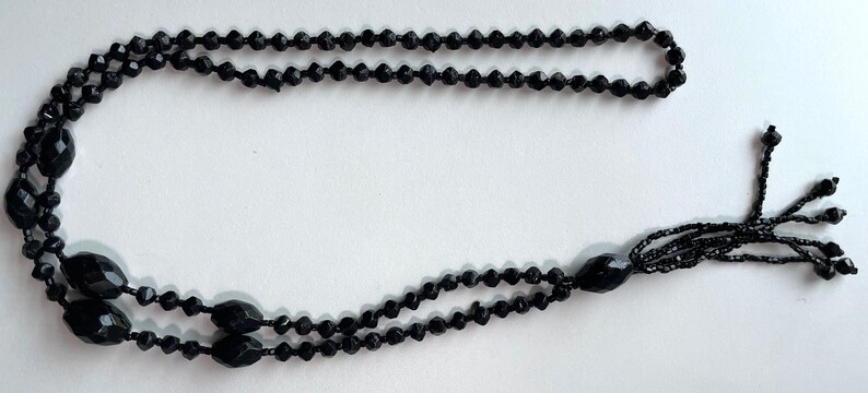 K&ouml;nnte beinhalten: Eine schwarze Perlenkette mit Quaste. Die Kette besteht aus kleinen, facettierten Perlen und hat eine lange, herabh&auml;ngende Quaste am Ende.