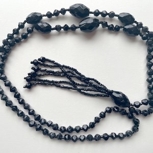 K&ouml;nnte beinhalten: Eine schwarze Perlenkette mit Quaste. Die Kette besteht aus facettierten Perlen und hat eine lange, baumelnde Quaste.