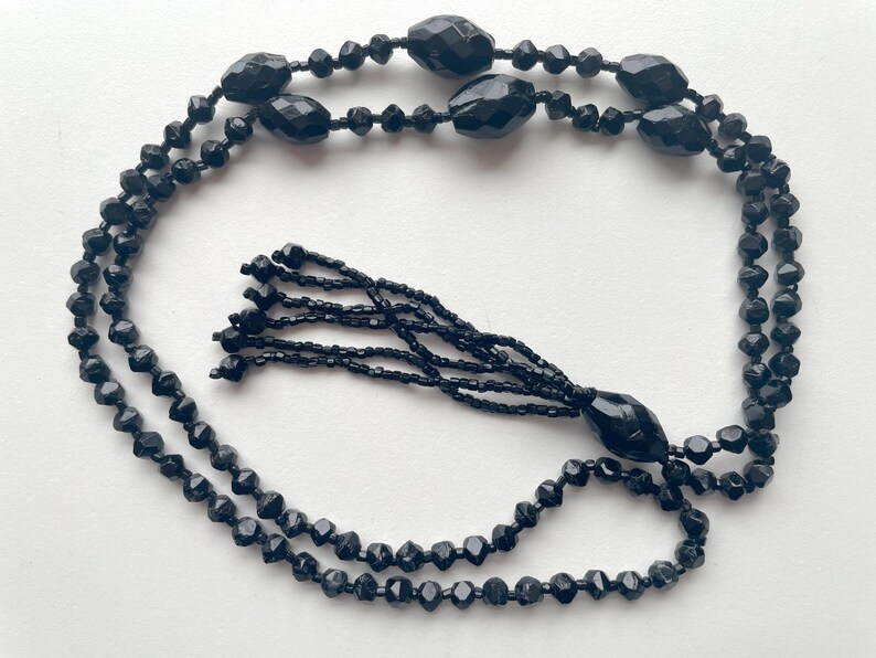 K&ouml;nnte beinhalten: Eine schwarze Perlenkette mit Quaste. Die Kette besteht aus facettierten Perlen und hat eine lange, baumelnde Quaste.