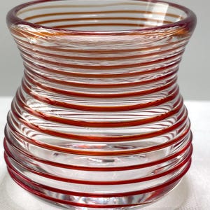 May include: A clear glass vase with horizontal red stripes. The vase has a rounded base, a slightly flared top, and a cylindrical body with a series of red bands. The glass appears to be hand-blown, with slight variations in the stripe width.