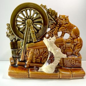May include: A ceramic figurine depicting a spinning wheel, a cat, and a dog. The figurine is primarily brown and tan with green accents. The dog is white and brown. The wheel is a prominent feature.