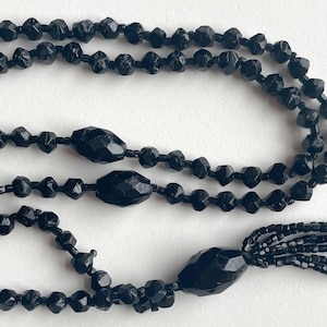 K&ouml;nnte beinhalten: Eine lange schwarze Perlenkette mit Quaste. Die Kette besteht aus facettierten Perlen und hat eine lange, herabh&auml;ngende Quaste am Ende.