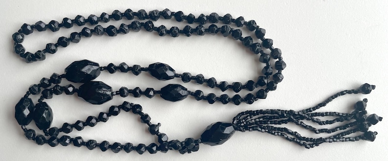 K&ouml;nnte beinhalten: Eine lange schwarze Perlenkette mit Quaste. Die Kette besteht aus facettierten Perlen und hat eine lange, herabh&auml;ngende Quaste am Ende.