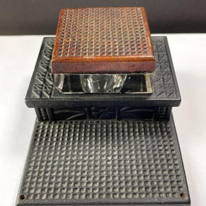 May include: A black inkwell with a wooden lid and a glass insert. The inkwell is decorated with a geometric pattern.