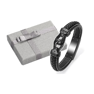 Könnte beinhalten: Schwarzes Lederarmband mit einem schwarzen Metallverschluss. Das Armband befindet sich in einer grauen Geschenkbox mit einem grauen Band.
