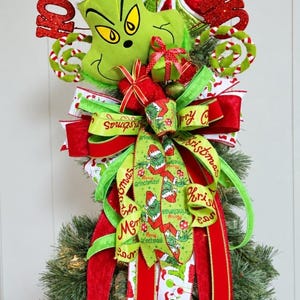 Op de afbeelding: Een kerstboompiek met een pluche groen karakterhoofd met een rode kerstmuts, omringd door rode glitterende HO-tekst, een groen-wit gestreept snoepornament en meerdere linten in rood, limoengroen en wit met bedrukte Grinch-afbeeldingen.