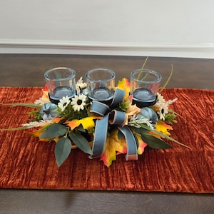 Puede incluir: Un centro de mesa de temática otoñal con tres candelabros de vidrio, calabazas falsas y hojas de otoño. El centro de mesa está sobre un camino de mesa de color óxido.