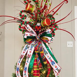 Op de afbeelding: Een kerstboompiek met een centrale notenkrakerfiguur, rood-groene trommels en rood-wit gestreepte linten. Het arrangement bevat lange rode glittertakken, groene bessenaccenten en een meerlaagse strik met ruitpatronen en notenkrakerafbeeldingen.