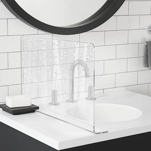 May include: A clear acrylic sink splash guard with water droplet details. The guard is positioned on a white countertop, next to a faucet and a white sink. A black soap dish with a white bar of soap is on the counter.