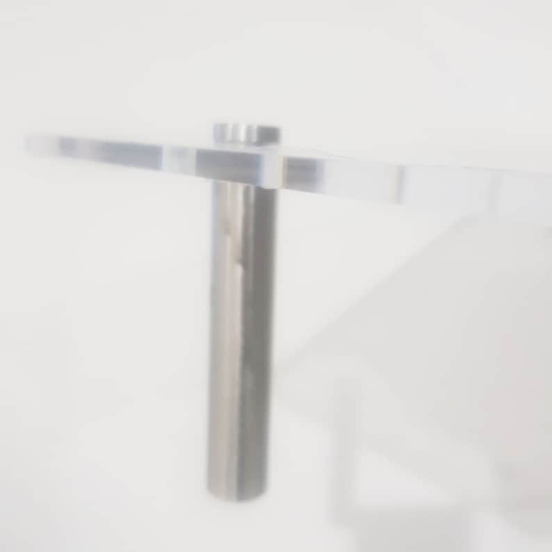 May include: A clear acrylic shelf with a silver metal bracket. The shelf is mounted to a white wall.