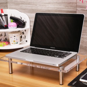 May include: A clear acrylic laptop stand with silver metal legs. The stand is on a wooden desk with a laptop on top of it.