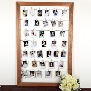 May include: A wooden photo display board with a brown frame and white backing. Small photos are clipped to horizontal strings with wooden pegs. A bouquet of white roses sits at the bottom right. The photos appear to be of a baby.