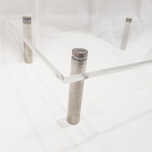 May include: A clear acrylic square display stand with four silver metal legs. The stand is made of clear acrylic and has a smooth, polished finish.
