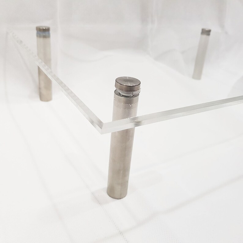 May include: A clear acrylic square display stand with four silver metal legs. The stand is made of clear acrylic and has a smooth, polished finish.