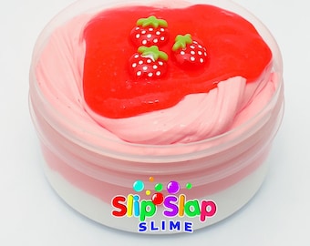 Puede incluir: Un recipiente de plástico transparente lleno de slime rosa y rojo con un remolino de fresa y tres encantos de fresa rojos en la parte superior. El recipiente tiene una etiqueta blanca con el texto "Slip Slap Slime" en él.