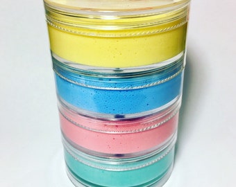 Puede incluir: Una pila de cuatro recipientes transparentes llenos de slime de diferentes colores. El slime es amarillo, azul, rosa y turquesa. El recipiente superior tiene una etiqueta que dice "Slap Slap Slime Butter Slime".