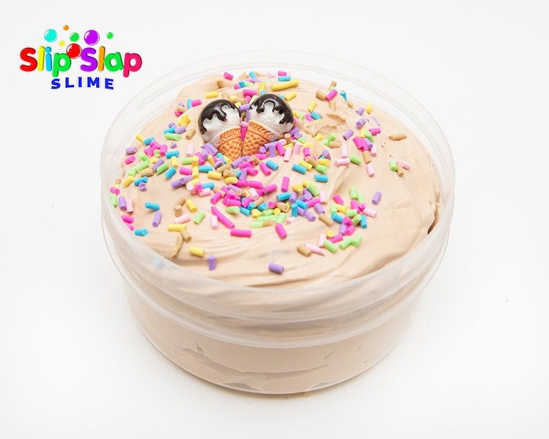 Puede incluir: Un recipiente de pl&aacute;stico transparente lleno de slime marr&oacute;n claro cubierto con chispas de colores y dos conos de helado en miniatura. El recipiente tiene una etiqueta blanca con el texto "Slip Slap Slime".