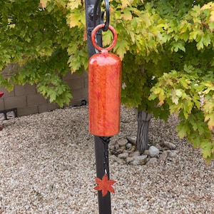 May include: A cylindrical wind chime featuring a vibrant orange and red marbled finish. The chime hangs from a black metal hook by a circular ring and includes a black chain with a star-shaped orange and red pendant at the bottom.