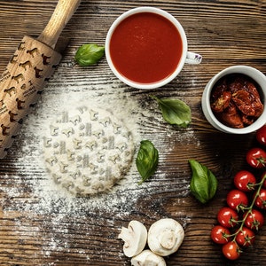 Op de afbeelding: Een houten deegroller met een bruine handgreep en een ontwerp van honden op het rolvlak. De deegroller ligt op een houten oppervlak met een deegbal, tomaten, basilicum en champignons.