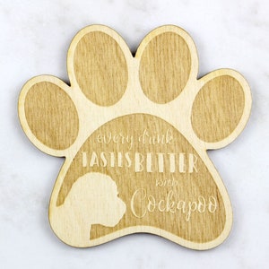 May include: Wooden paw-shaped coaster with the text "every drink tastes better with Cockapoo" and a Cockapoo silhouette. The coaster is made of light-colored wood and has a natural wood grain finish.