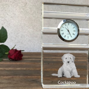 May include: A clear glass clock with a white face and black Roman numeral hour markers. The clock is sitting on a clear glass base with a white dog etched into the glass. The dog is sitting and the text "Cockapoo" is below the dog.