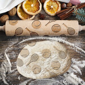 May include: A wooden rolling pin with a tennis ball and racket design. The rolling pin is on a wooden surface with flour scattered around it.