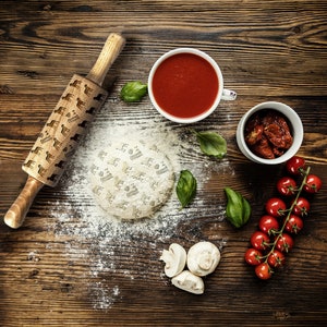 Op de afbeelding: Een houten deegroller met een patroon van honden erop, een kom met tomatensaus, een kom met gedroogde tomaten, een kom met cherrytomaatjes, een pizzabodem met een hondenpatroon en verse basilicumblaadjes op een houten oppervlak.
