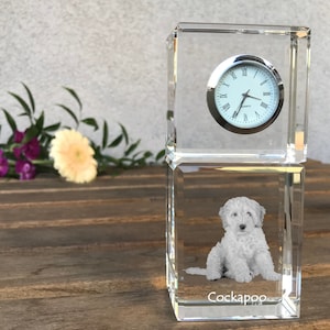 May include: A crystal clock with a white clock face and black hands. The clock is on a clear crystal base with a white Cockapoo dog engraved inside. The base is sitting on a wooden surface.