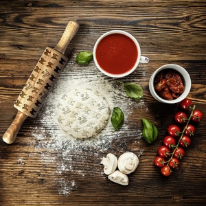 Op de afbeelding: Een houten deegroller met een vlinderontwerp ligt op een houten tafel met pizzadough, tomatensaus, cherrytomaatjes, basilicum en champignons.