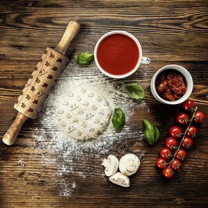 Può includere: Un mattarello di legno con un motivo a forma di cuore viene utilizzato per stendere la pasta per pizza su una superficie di legno. L'impasto è ricoperto di farina. Ci sono altri ingredienti per la pizza sulla superficie, tra cui salsa di pomodoro, pomodorini, pomodori secchi e funghi.
