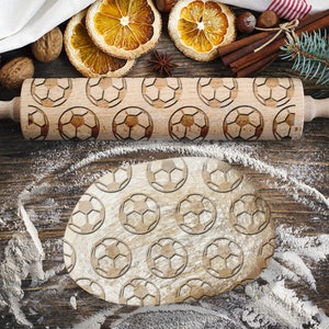 May include: A wooden rolling pin with a soccer ball design. The rolling pin is on a wooden surface with flour scattered around it.