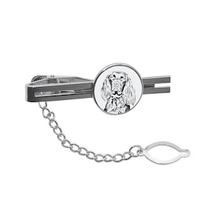 May include: Silver tie clip with a round, white enamel charm featuring a black and white illustration of a dog's head. The tie clip has a chain attached to a small, silver loop.