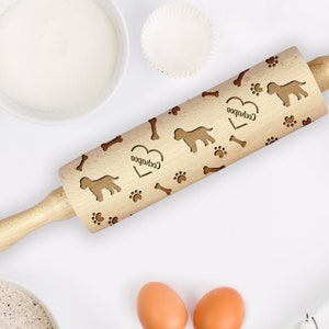Puede incluir: Un rodillo de madera con un corazón y la palabra "Cockapoo" grabada. El rodillo está decorado con un patrón repetitivo de huellas de patas, huesos y siluetas de Cockapoo.